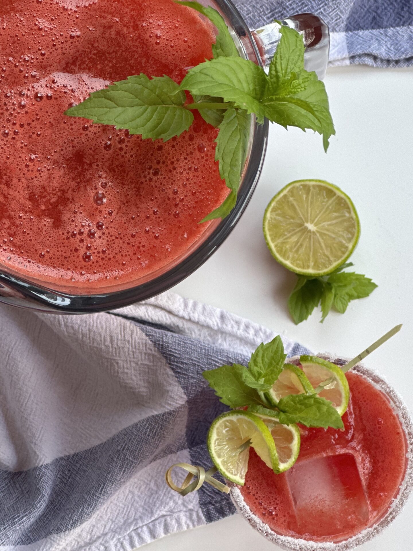 A glass and a pitcher of freshly made watermelon agua fresca are seen from above, surrounded by a blue checkered cloth and fresh mint and lime