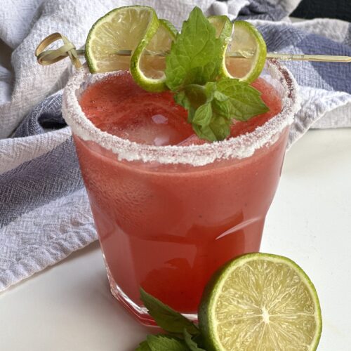 A sugar rimmed glass of watermelon agua fresca garnished with fresh mint and lime sits on a white table.