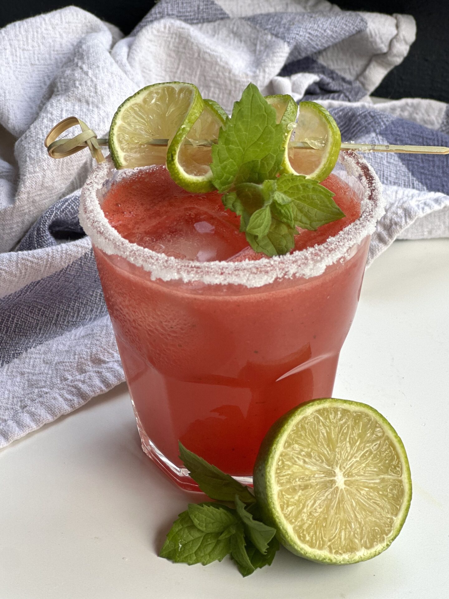 A sugar rimmed glass of watermelon agua fresca garnished with fresh mint and lime sits on a white table.