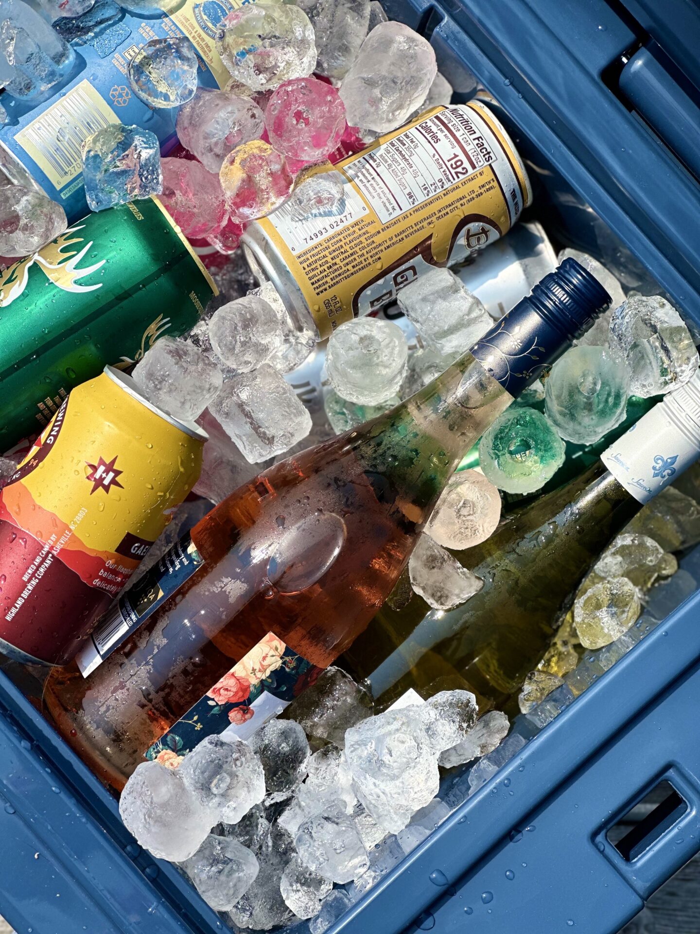 An open dark blue cooler filled with  ice, drink cans and wine bottles is seen from above