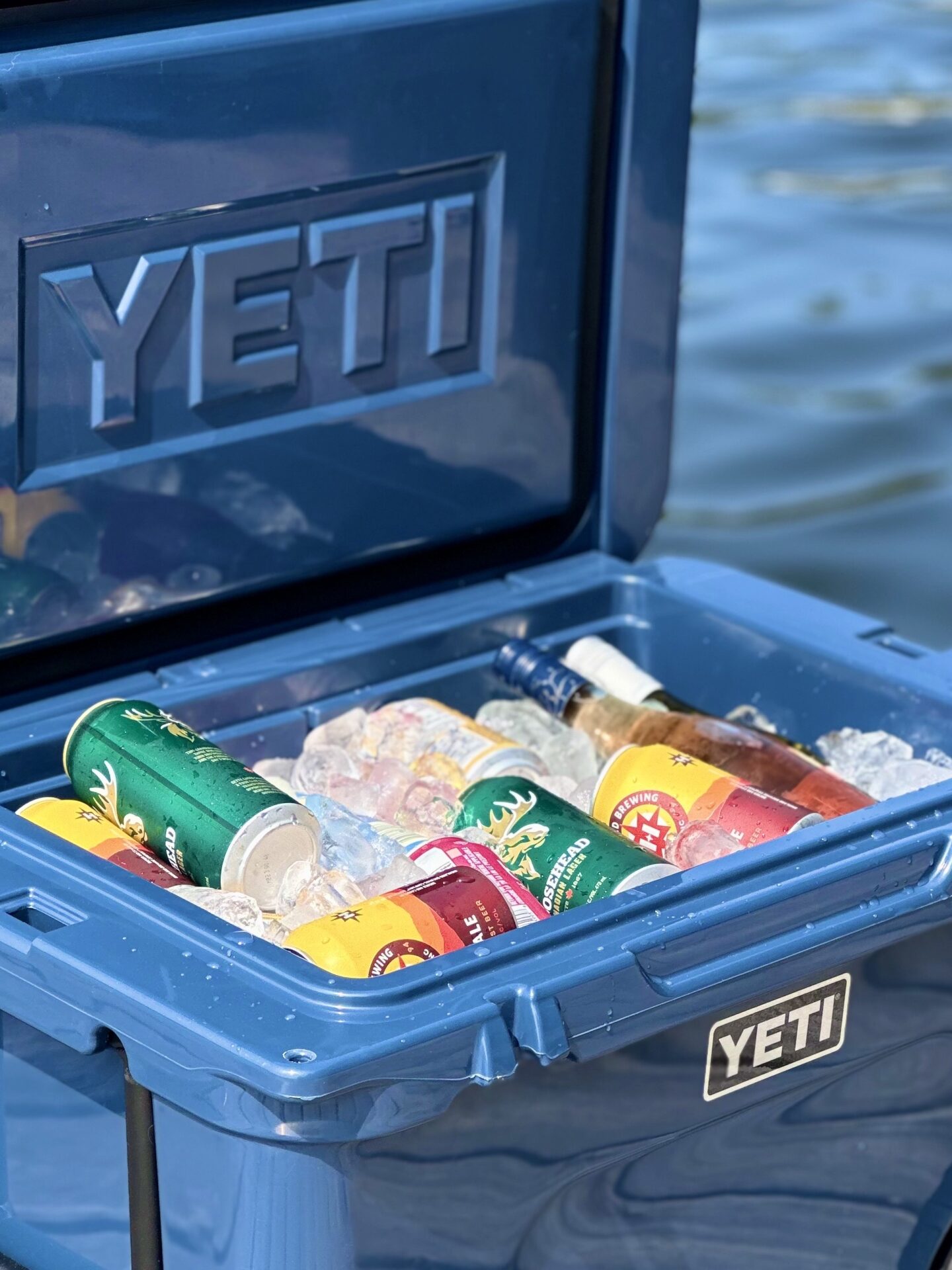 A blue Yeti cooler filled with ice and cold drinks sits open on a dock by a lake