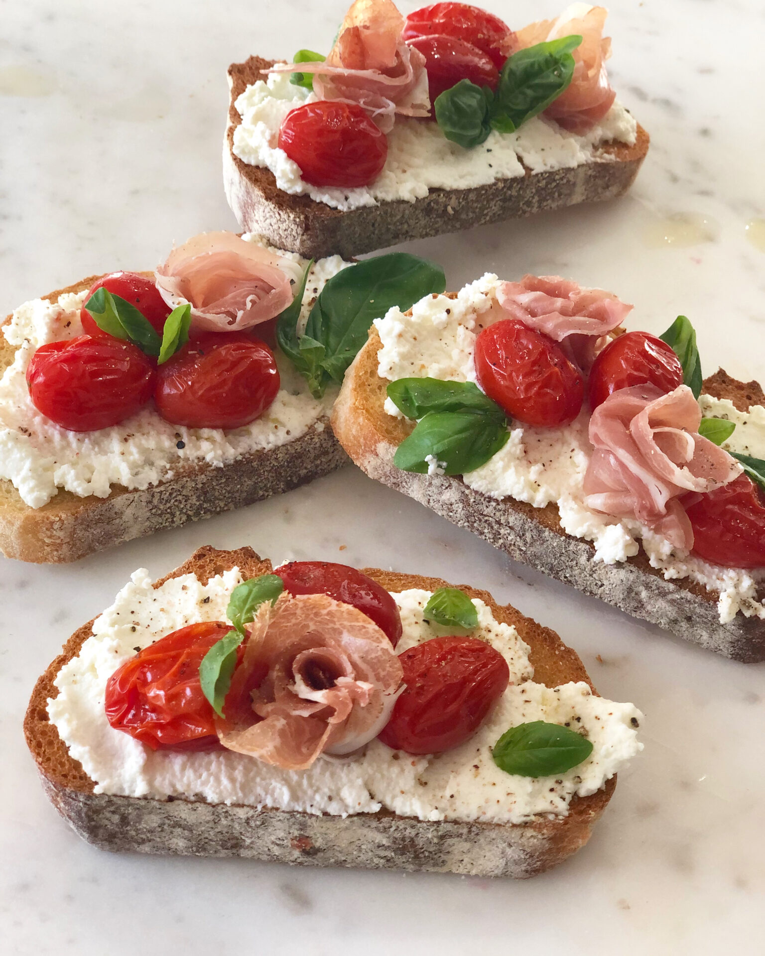 Thick slices of toast are seen slathered in homemade ricotta cheese and topped with sliced cherry tomatoes, fresh basil and prosciutto rosettes