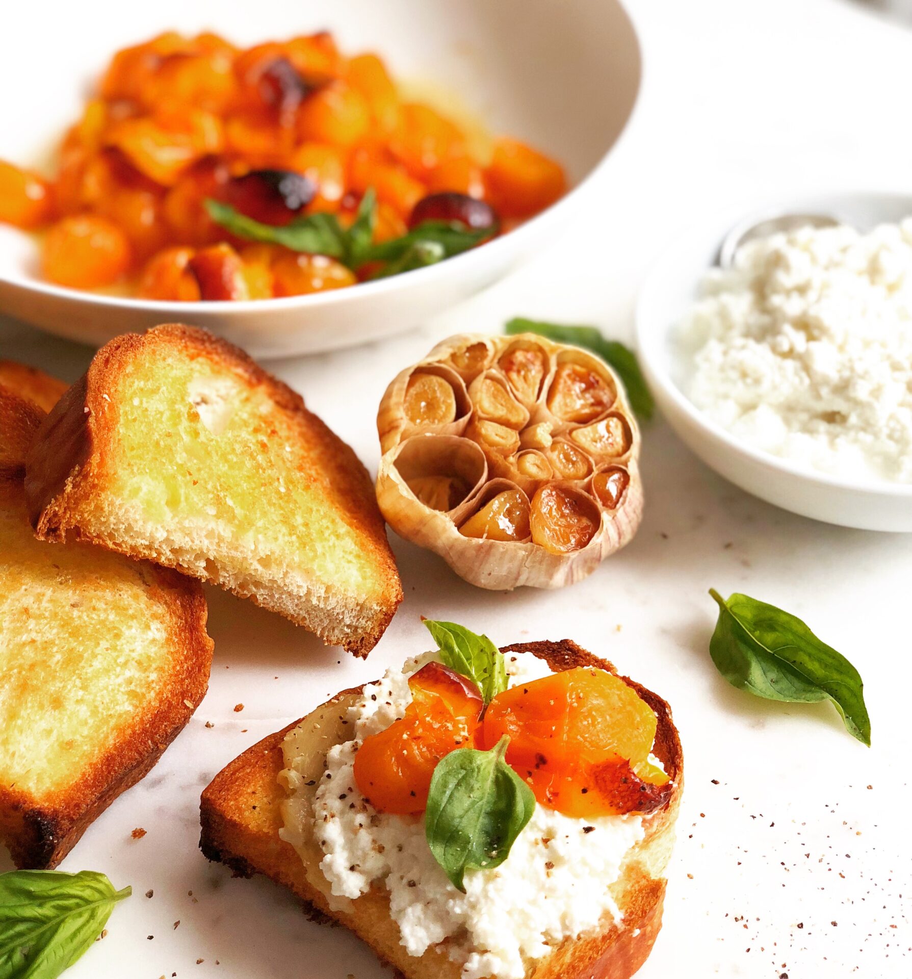 Garlic bread is seen slathered with ricotta cheese and topped with roasted yellow cherry tomatoes and fresh basil