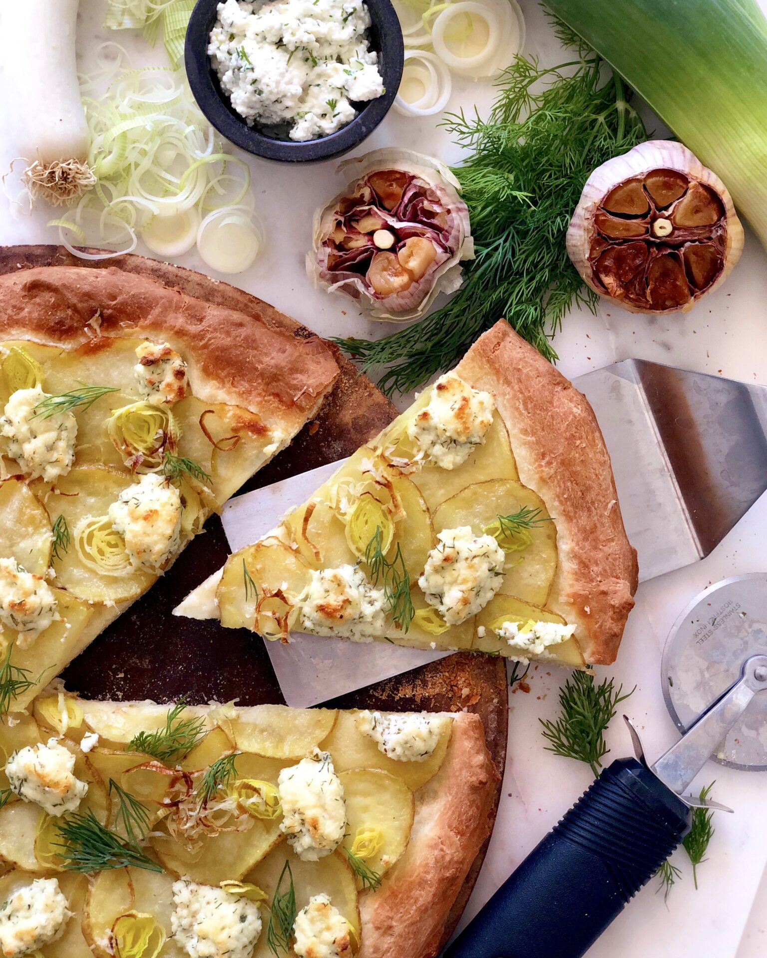 A potato, leek and ricotta pizza is seen from above, surrounded by heads of roasted garlic and fresh herbs