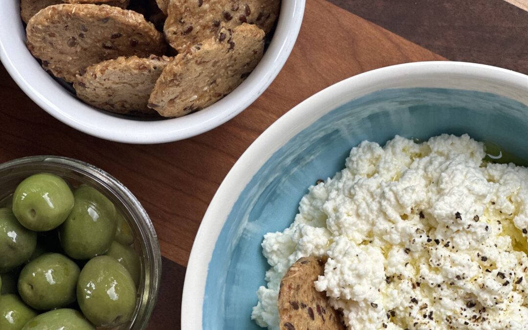 A bowl of homemade ricotta cheese is seen from above, surrounded by crisp crackers and bright green olives