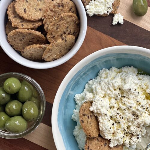 A bowl of homemade ricotta cheese is seen from above, surrounded by crisp crackers and bright green olives