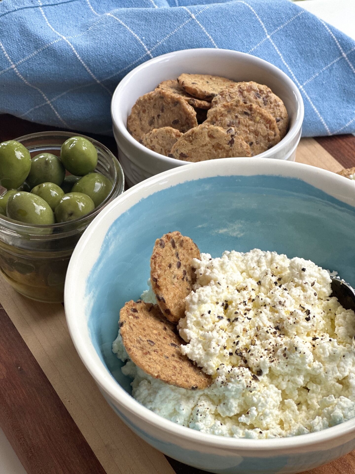 A bowl of homemade ricotta cheese is seen on a serving board with crackers and olives