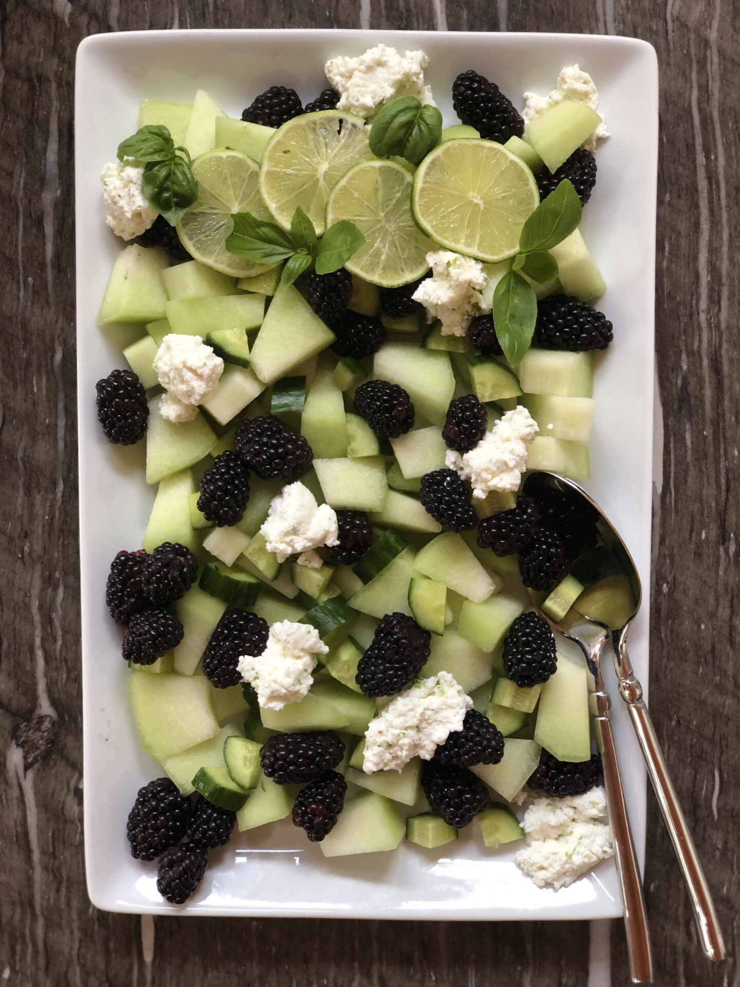 A salad of honeydew melon, blackberries, cucumbers and ricotta cheese is seen from above, garnished with fresh mint and lime wheels