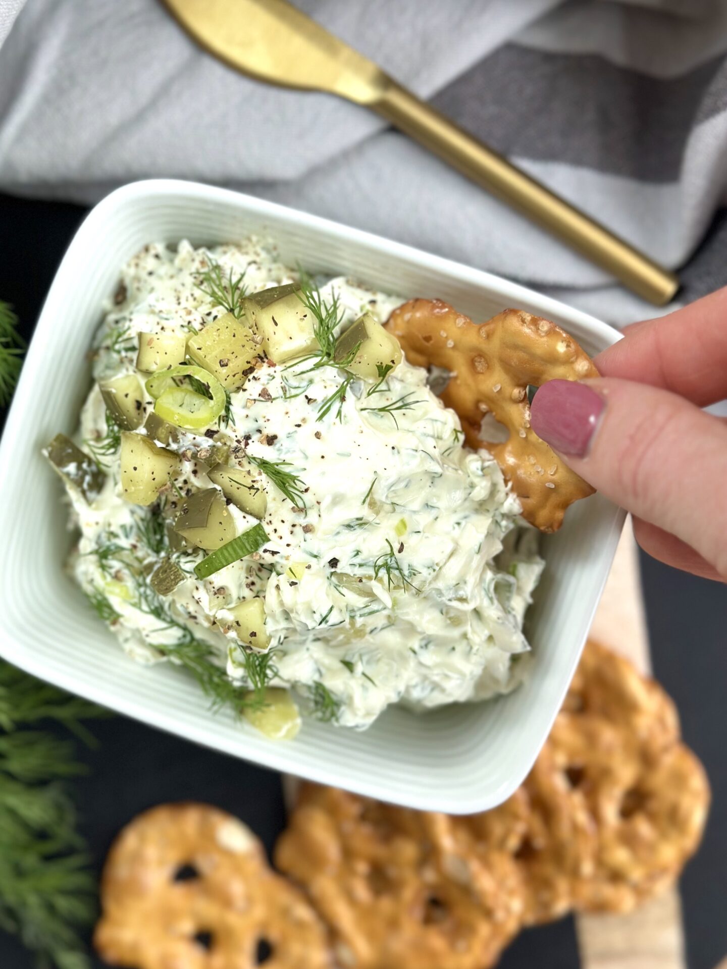 A woman's hand is seen dipping a pretzel chip into a bowl of dill pickle dip garnished with chopped dill pickles and fresh dill.