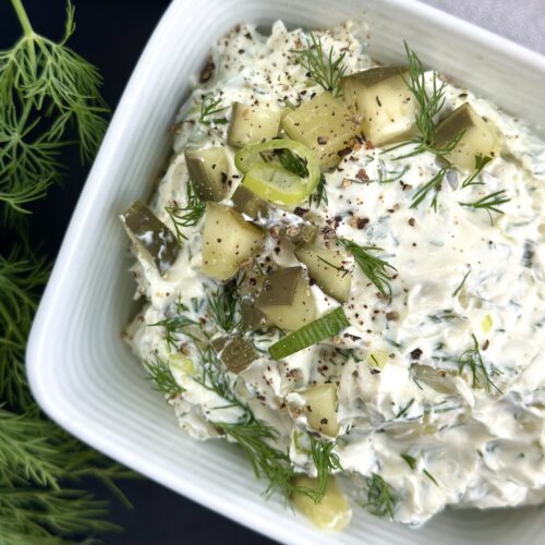 A square dish of dill pickle dip is seen close up, from above, garnished with chopped dill pickles, fresh dill, chopped green onions and freshly ground black pepper.