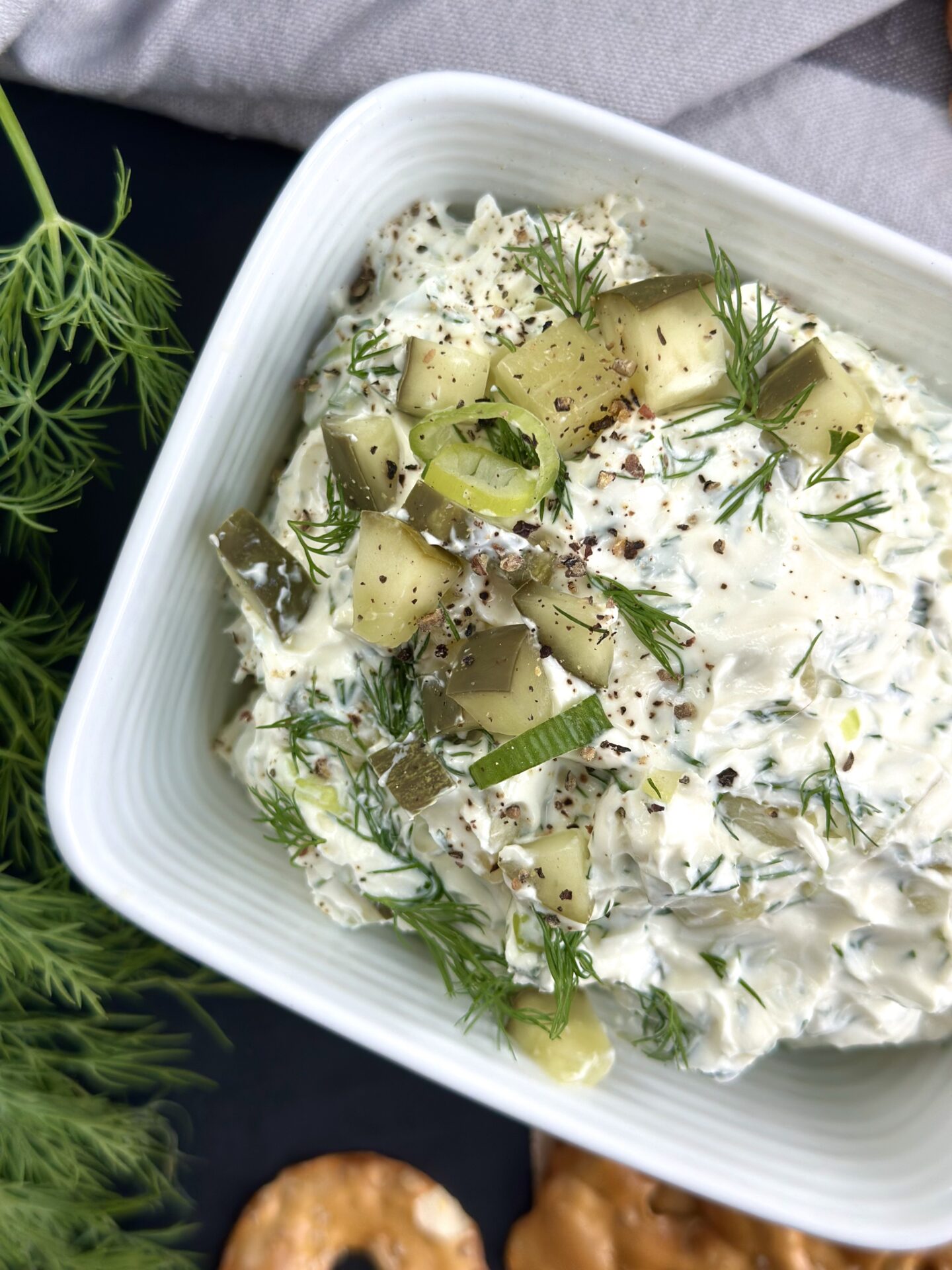 A square dish of dill pickle dip is seen close up, from above, garnished with chopped dill pickles, fresh dill, chopped green onions and freshly ground black pepper.