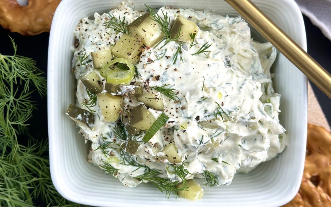 A bowl of dill pickle dip is seen from above, garnished with chopped pickles and surrounded by pretzel chips and fresh dill.