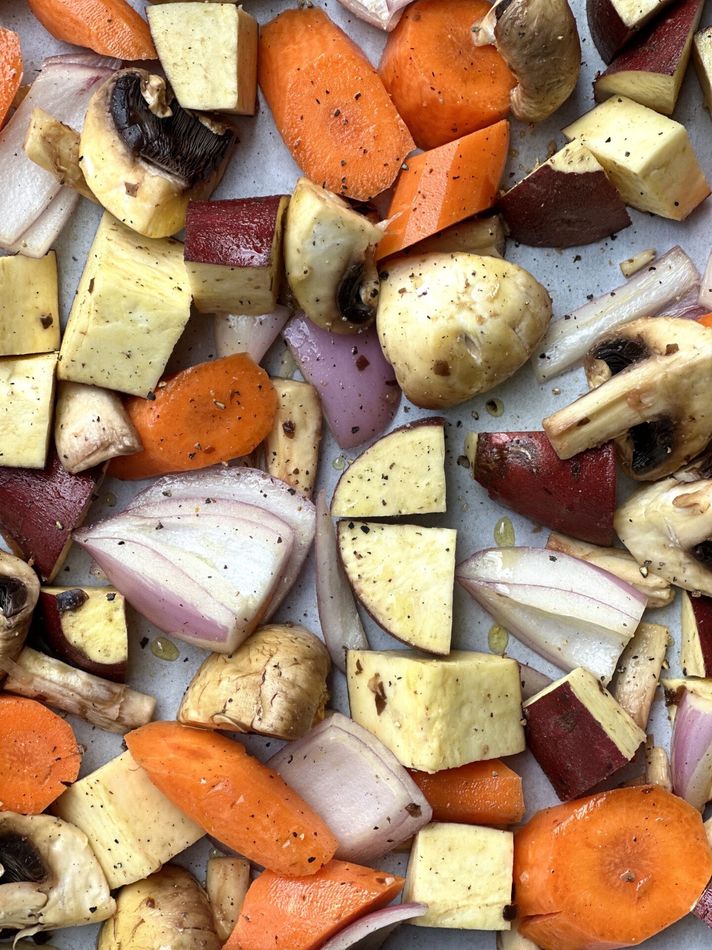 A medley of diced carrots, Japanese sweet potato, mushrooms and red onion are seen from above, dusted with salt and pepper and ready for roasting in the oven