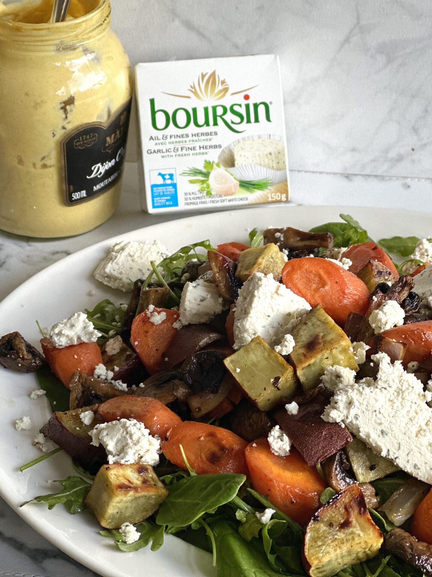 A platter of Warm Roasted Vegetable and Boursin Salad is seen from the side with a package of Boursin cheese and a jar of dijon mustard seen in the background.