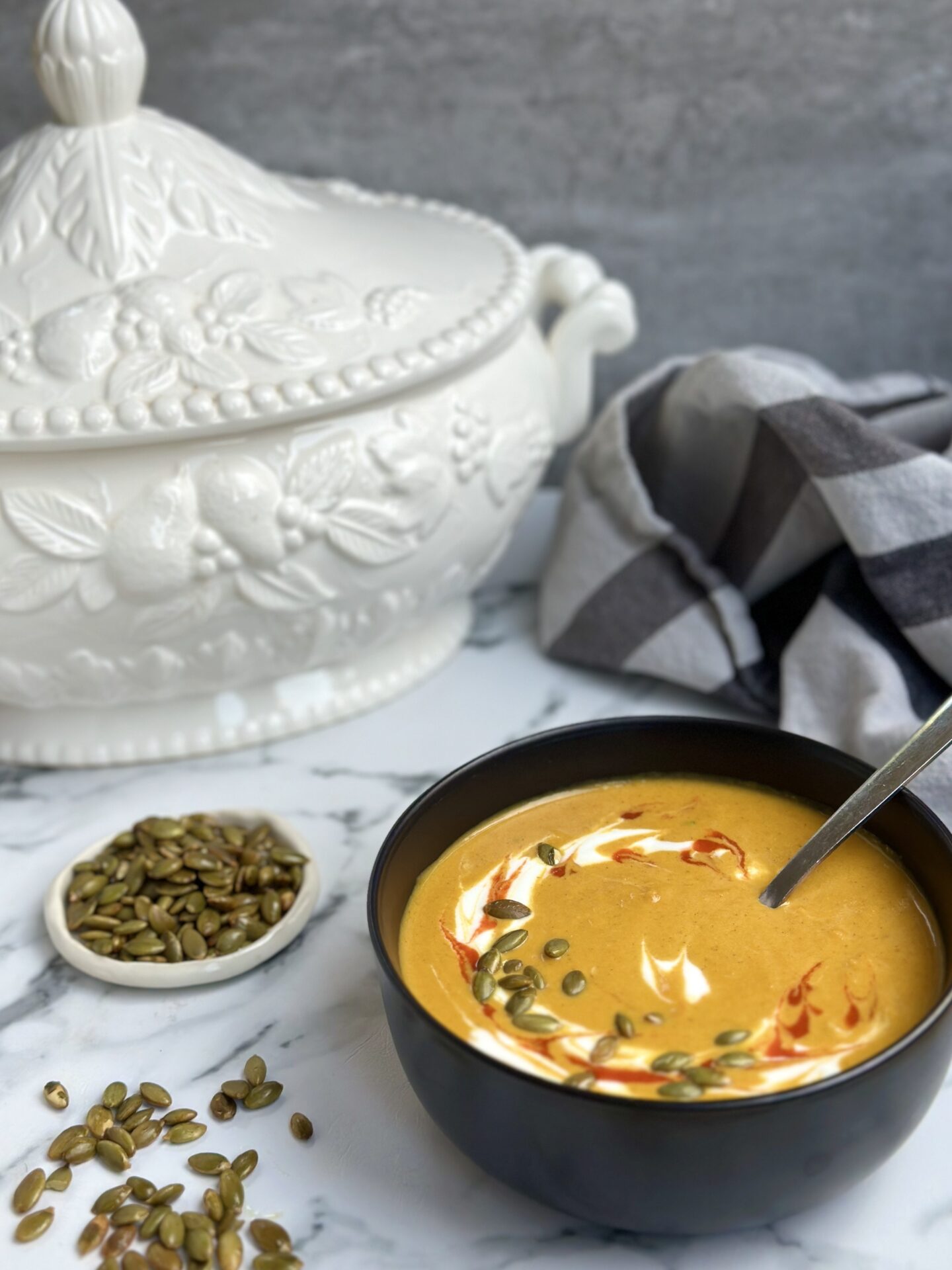 A black bowl of golden Creamy Pumpkin Spice Soup is seen on a white marble table surrounded by a a soup tureen and a small bowl of pumpkin seeds