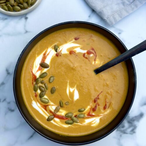 A black bowl filled with golden creamy pumpkin soup is seen from above garnished with swirls of yogurt and hot sauce and sprinkled with green pumpkin seeds