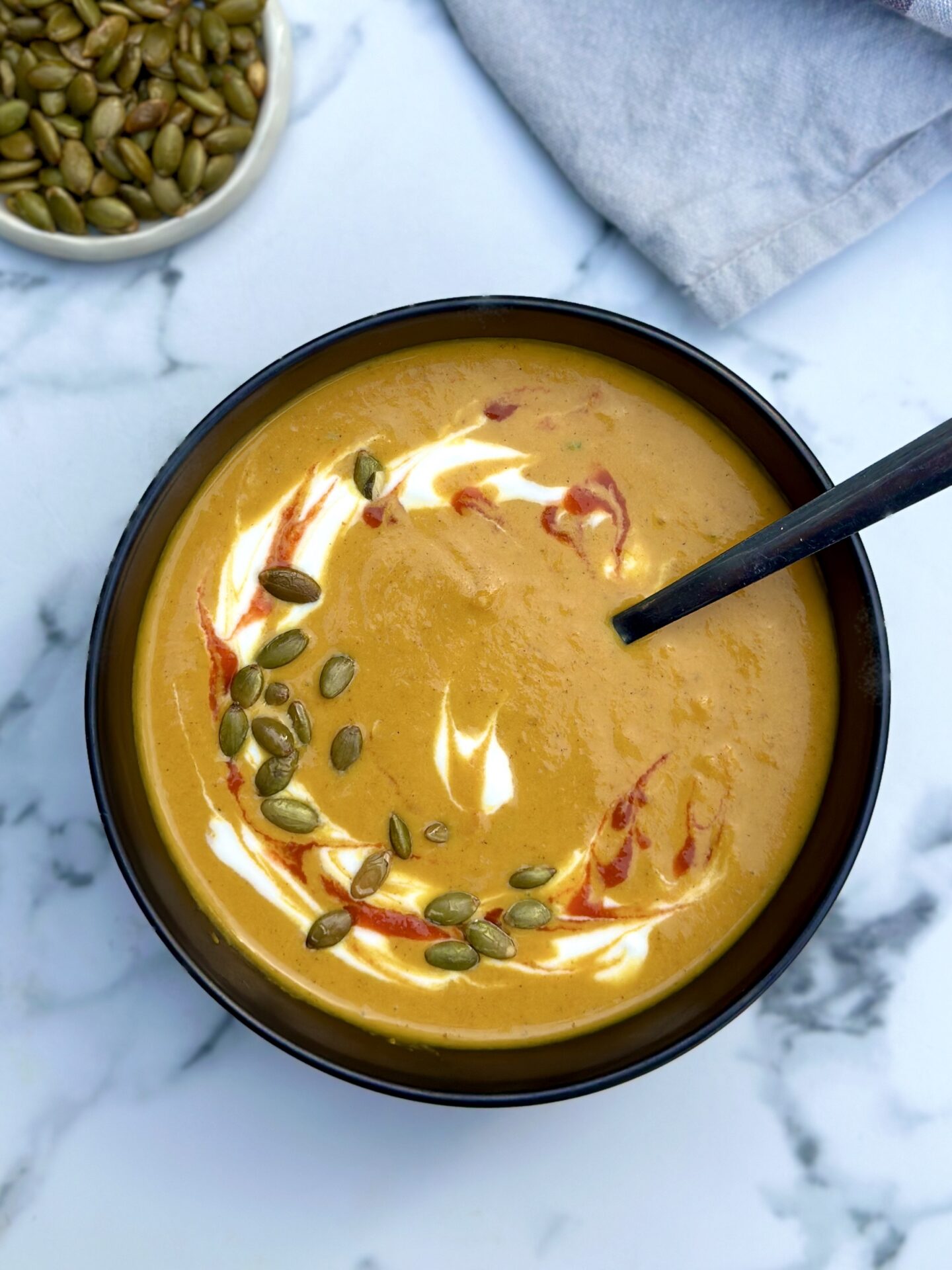 A black bowl filled with golden creamy pumpkin soup is seen from above garnished with swirls of yogurt and hot sauce and sprinkled with green pumpkin seeds