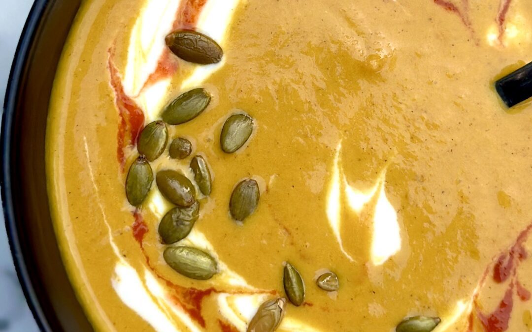 A black bowl filled with golden yellow Creamy Pumpkin Spice Soup is seen from above, garnished with swirls of white yogurt and red sriracha, and sprinkled with pumpkin seeds