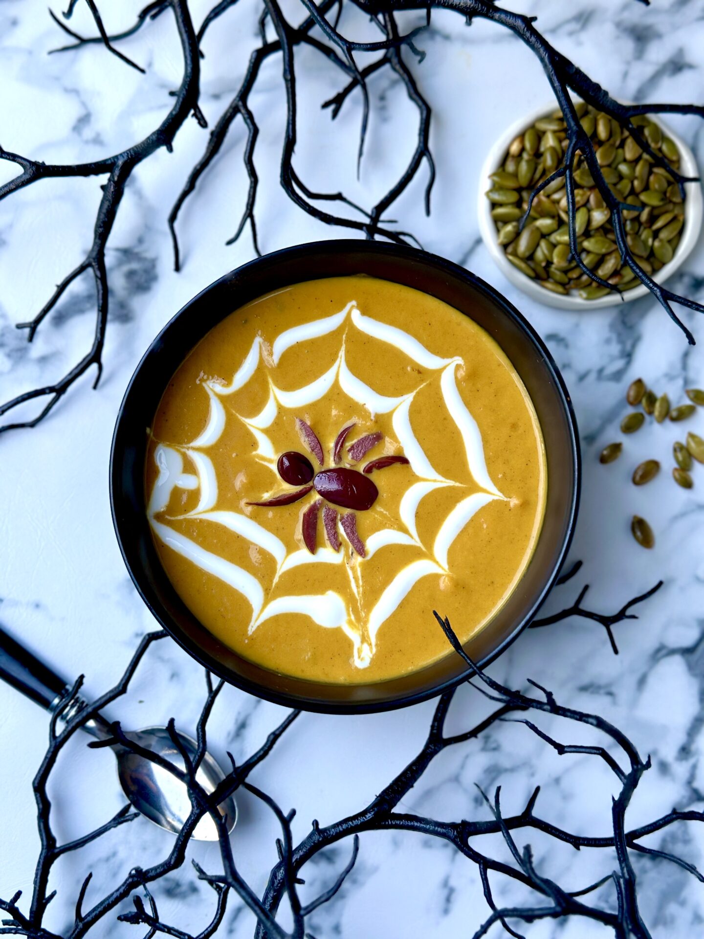 A black bowl of golden Creamy Pumpkin Soup is decked out for Halloween with a yogurt web and black olive spider garnish. The bowl rests on a black veined white marble table and is surrounded by black branches for a spooky effect.