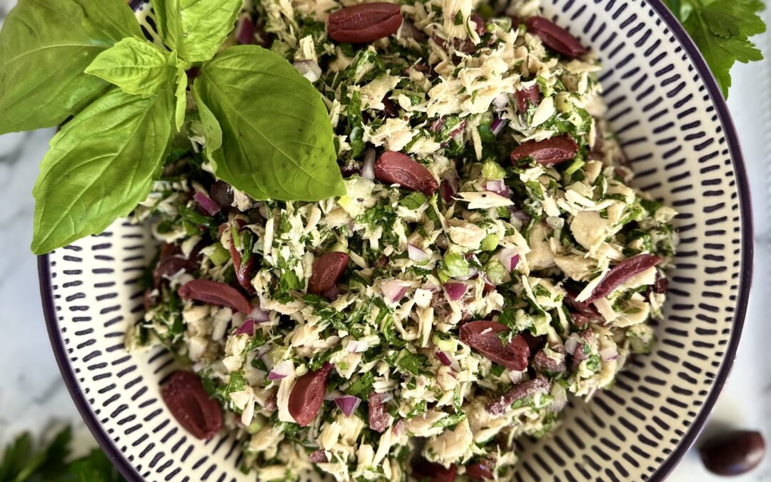 A bowl of Italian tuna salad is seen from above, garnished with fresh basil leaves. The bowl sits on a white marble table and is surrounded by parsley, lemons, red onions and black olives.