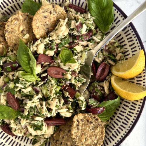 A bowl of Italian tuna salad is seen from above, surrounded by crackers, black olives and lemon wedges