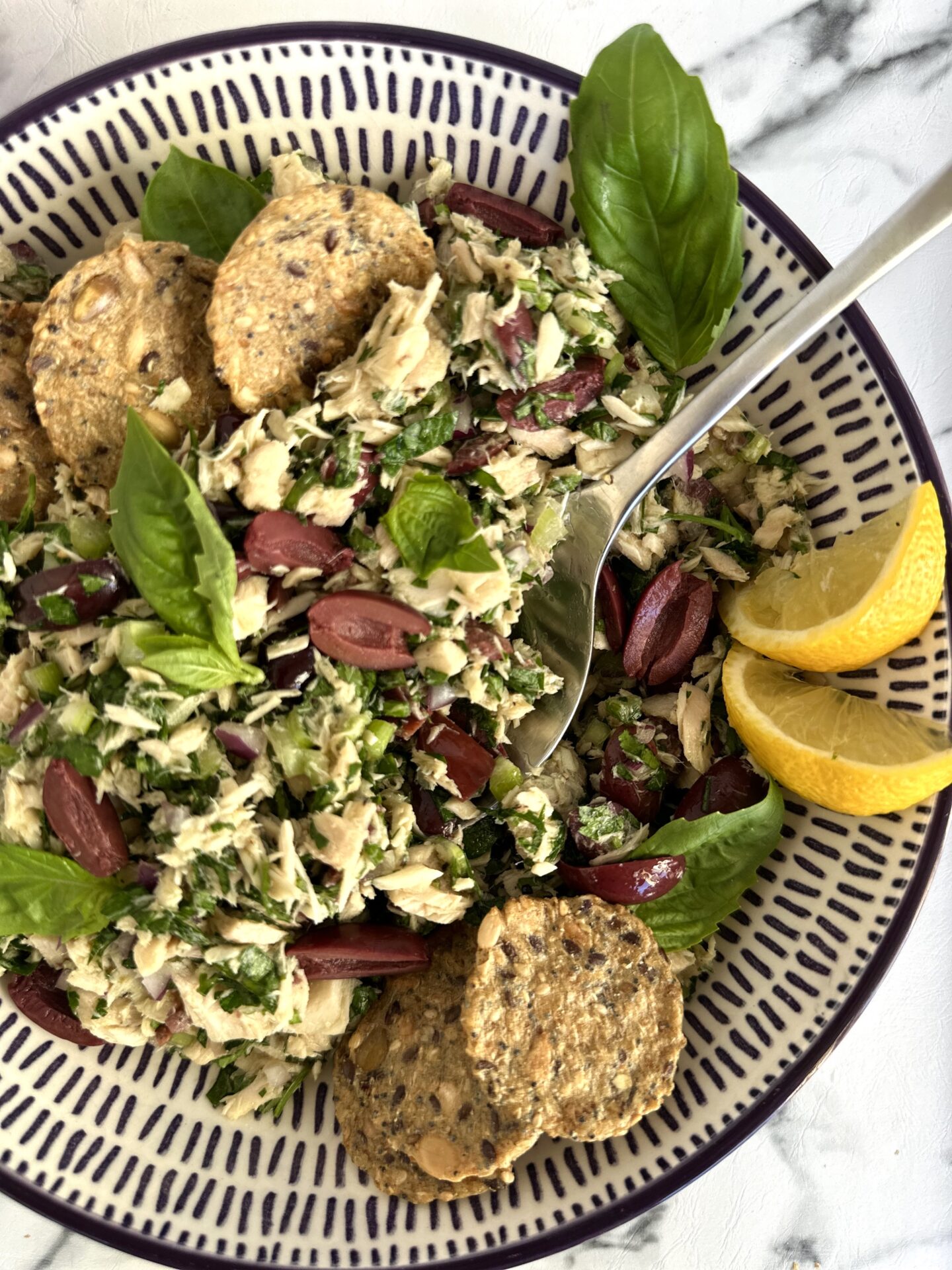 A bowl of Italian tuna salad is seen from above, surrounded by crackers, black olives and lemon wedges