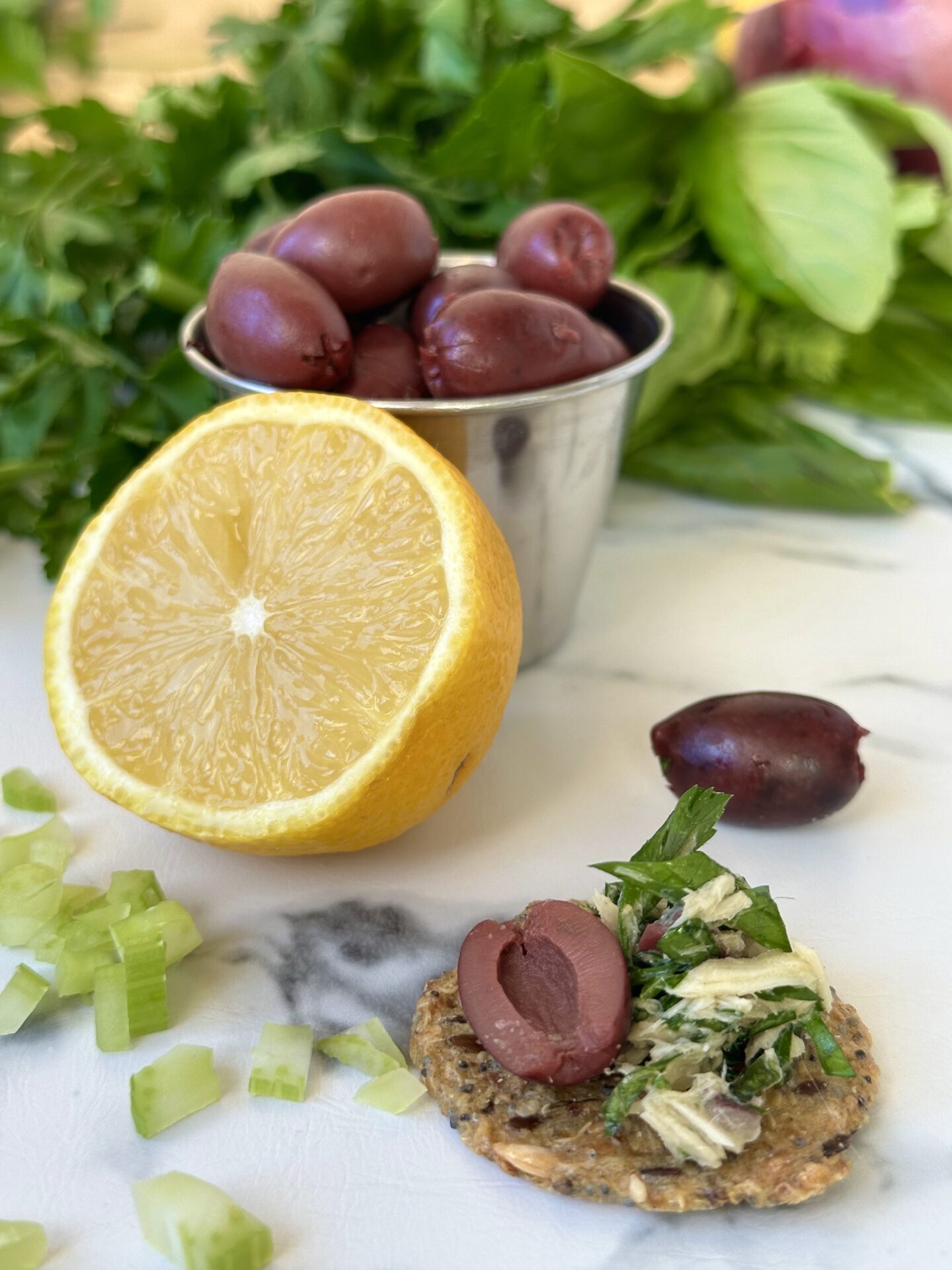 A cracker topped with Italian tuna salad sits on a white marble table, surrounded by lemons, black olives, chopped celery and lots of fresh green herbs.