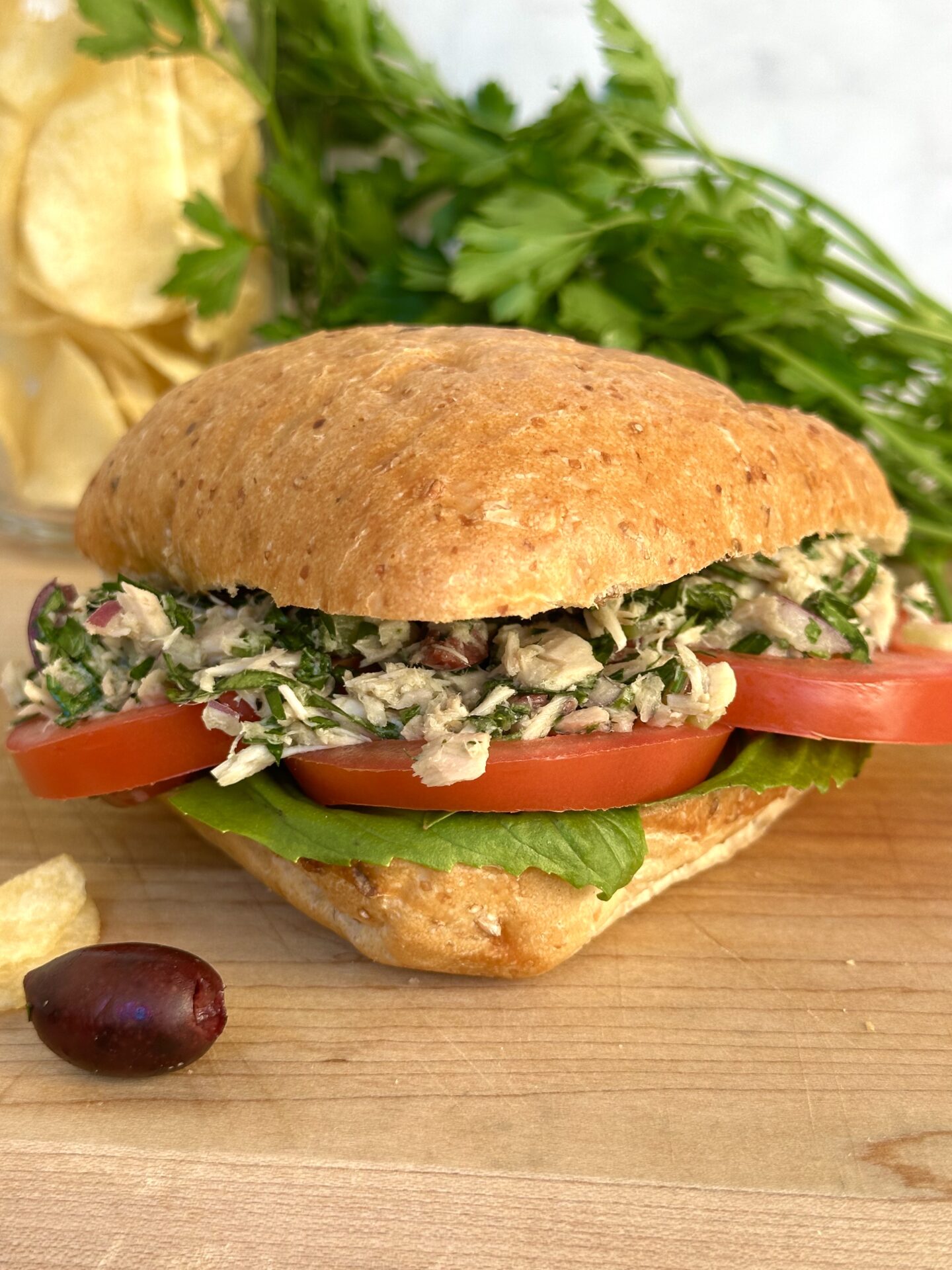 A large ciabatta bun is stuffed full of Italian tuna salad, thick red tomato slices and fresh basil leaves.  The sandwich sits on a wood serving board, surrounded by black olives, potato chips and parsley.