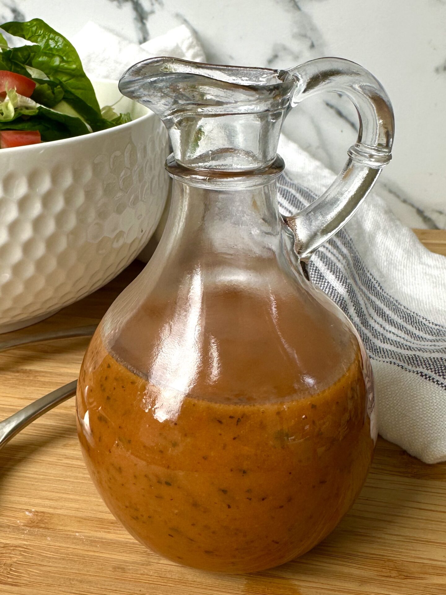 A glass cruet of homemade sundried tomato vinaigrette is seen close up, with a bowl of green salad and a white and blue striped tea towel in the background.