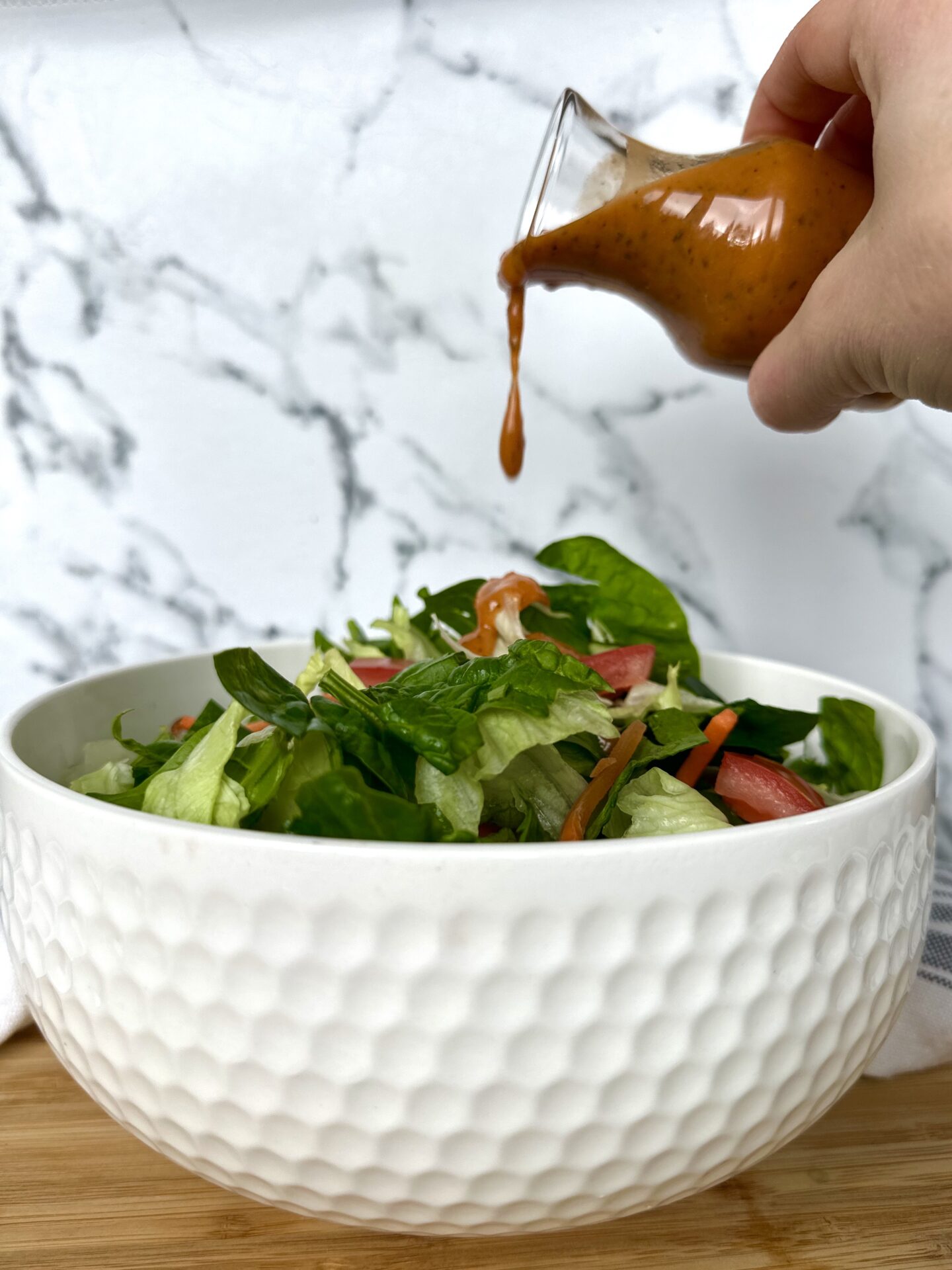 A hand pours Sundried Tomato Vinaigrette over a big white bowl filled with a green salad