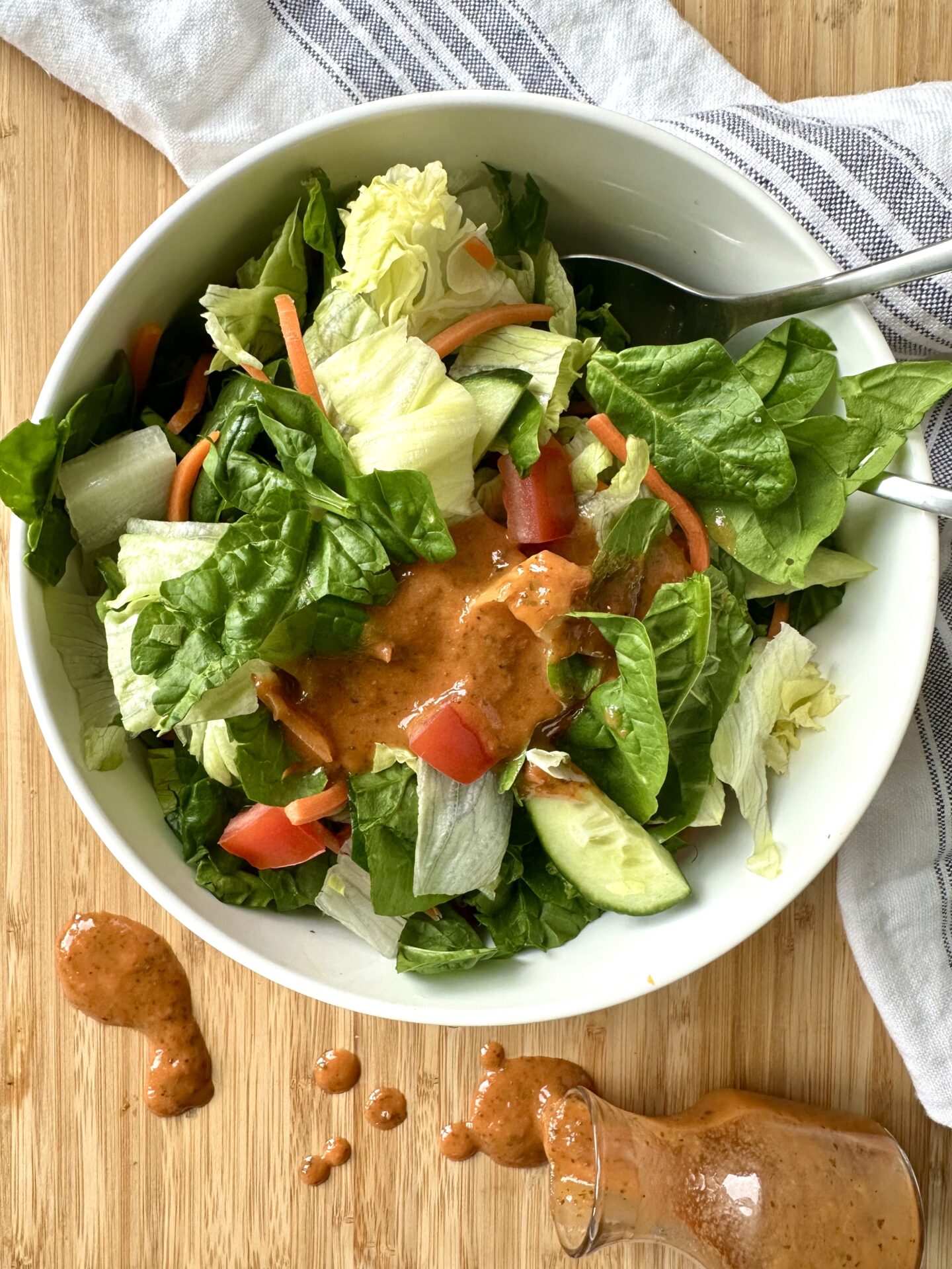 A bowl of garden salad is seen from above, drizzled with sundried tomato vinaigrette. The bottle of vinaigrette has been knocked over beside the salad and the dressing spills out in a pretty orange river onto the cutting board underneath