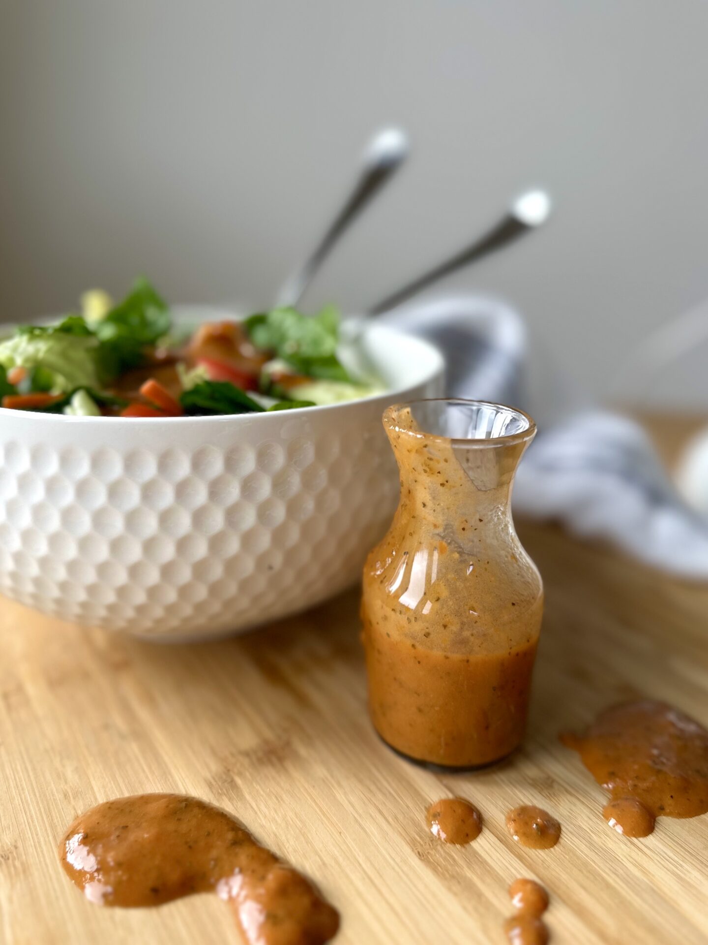 A white bowl filled with green salad sits on a wood chopping block next to a small glass bottle of sundried tomato vinaigrette. The vinaigrette has spilled, making puddles and drops of the dressing on the chopping board.