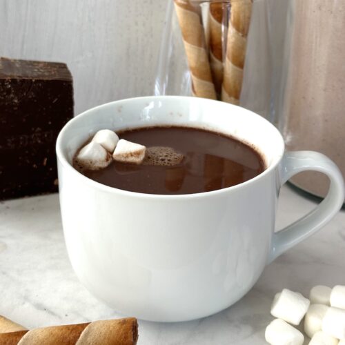 A large mug of rich dark brown hot chocolate is garnished with mini marshmallows and surrounded by cookie rolls and a big block of dark chocolate