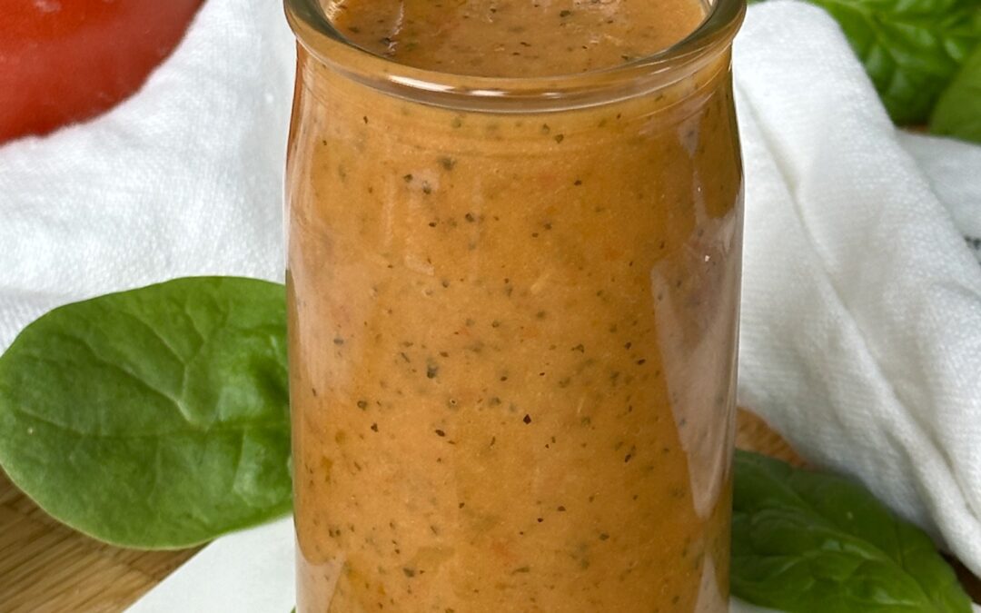 A jar of Sundried Tomato Vinaigrette sits on a wood table surrounded by bright red tomatoes and salad greens