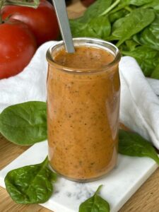 A jar of Sundried Tomato Vinaigrette sits on a wood table surrounded by bright red tomatoes and salad greens