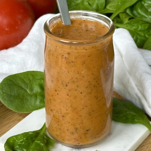 A jar of Sundried Tomato Vinaigrette sits on a wood table surrounded by bright red tomatoes and salad greens