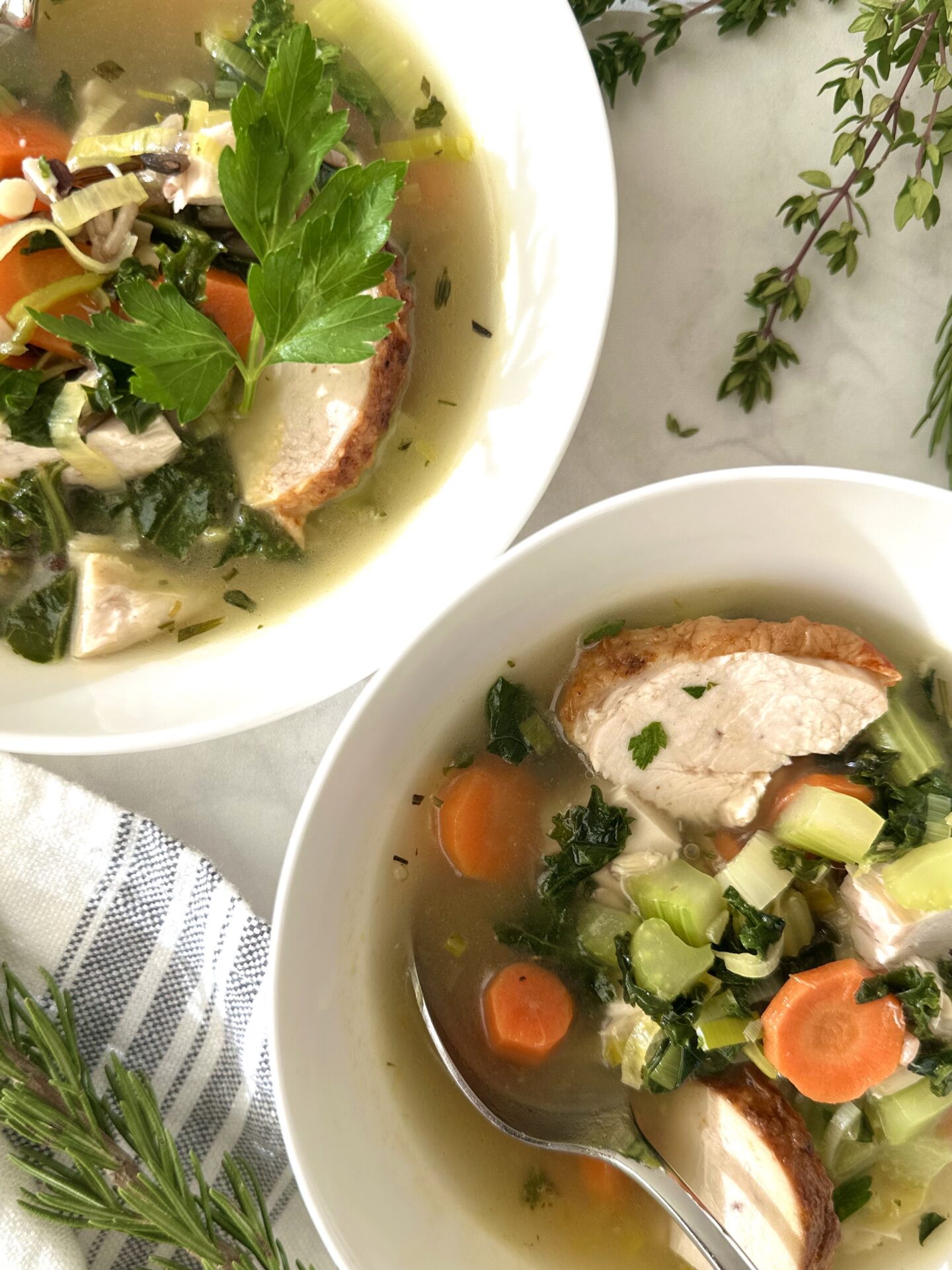 Two individual bowls of turkey, leek and wild rice soup sit on a white marble table surrounded by fresh herbs