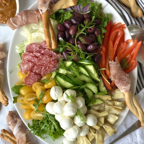 A platter of Antipasti Salad with Prosciutto Wrapped Bread Sticks is seen from above. The platter overflows with fresh, colourful vegetables, olives, salami and cheese and is surrounded by a bowl of sundried tomato dressing and prosciutto wrapped breadsticks.