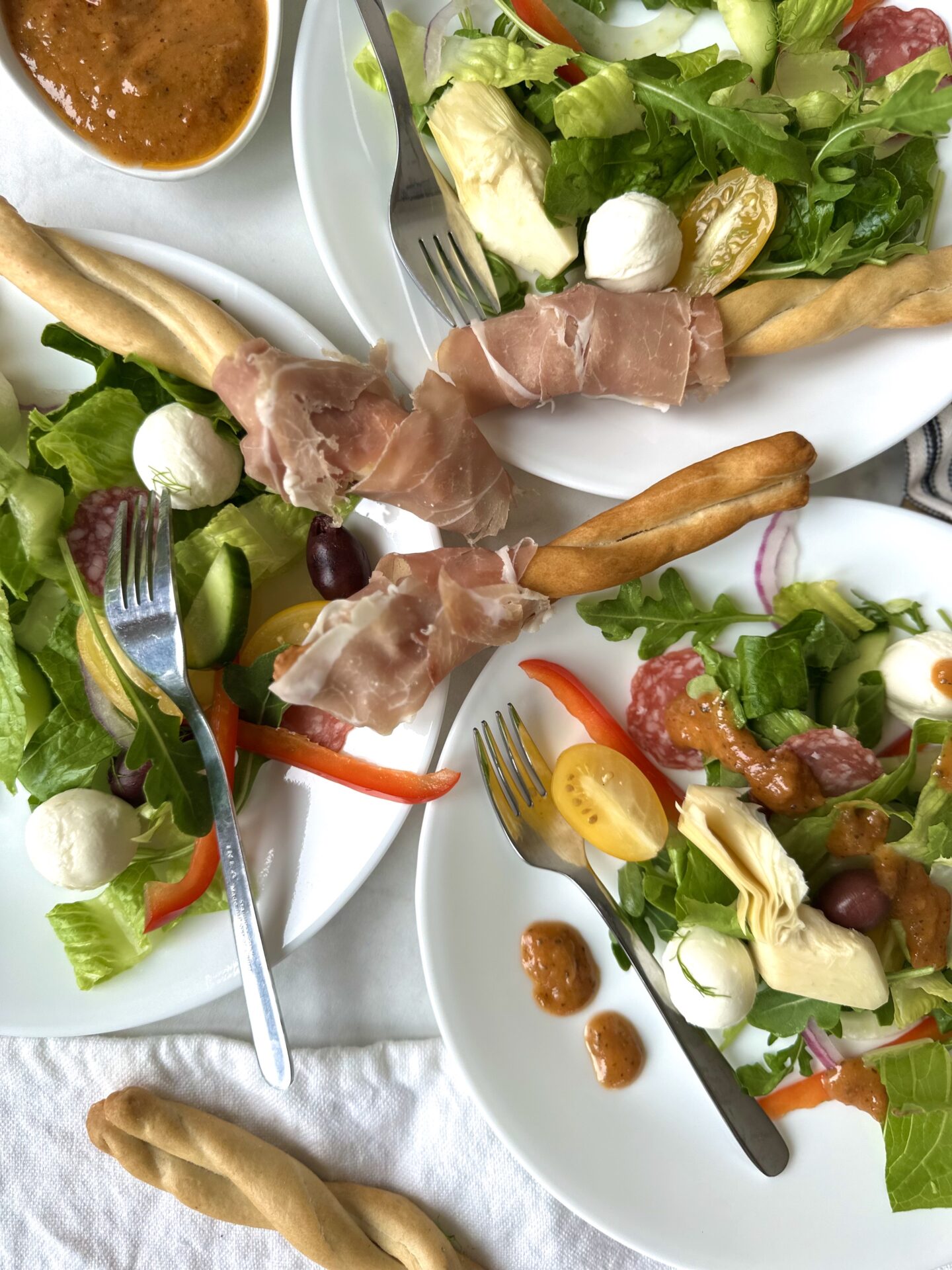 A trio of individual white plates is seen from above. Each plate has a personalized serving of Antipasti Salad with Prosciutto Wrapped Bread Sticks drizzled in Sundried Tomato Vinaigrette.