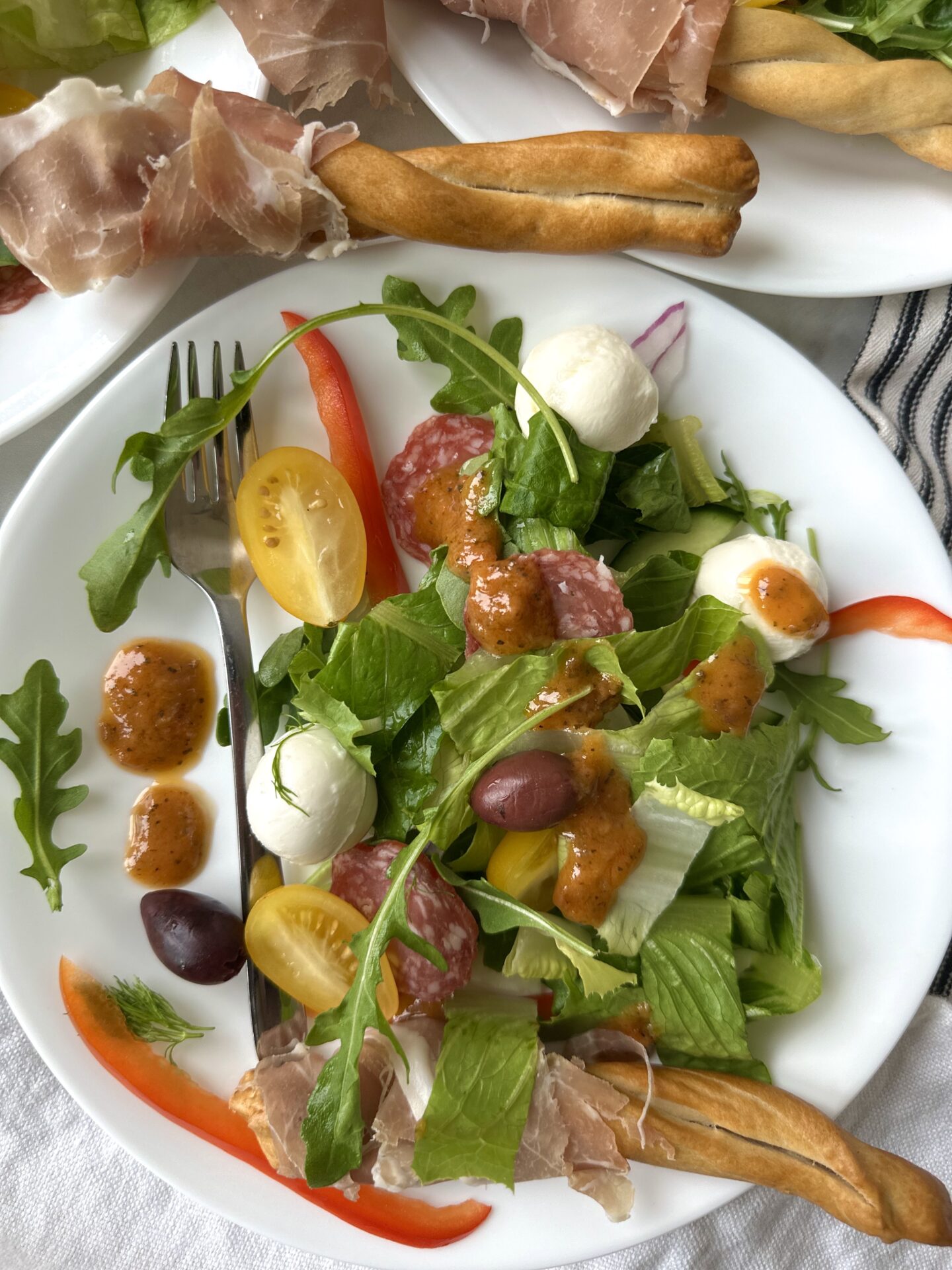 A single serving of Antipasti Salad with Prosciutto Wrapped Breadsticks is seen from above, drizzled in sundried tomato vinaigrette. More breadsticks can be seen in the background.