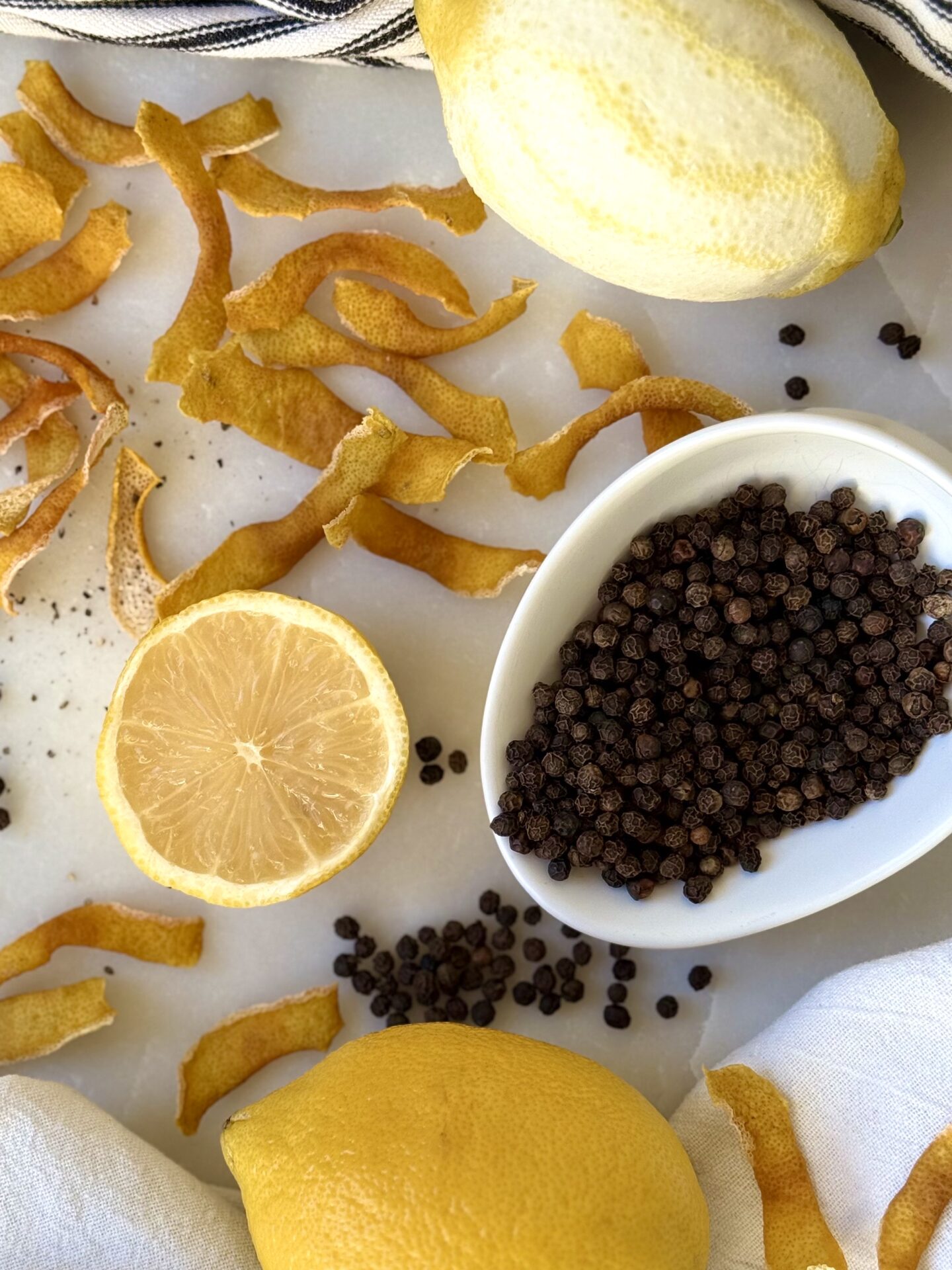 The ingredients for Homemade Lemon Pepper Seasoning - lemon zest, lemons and peppercorns -  are seen from above, on a white marble table.  