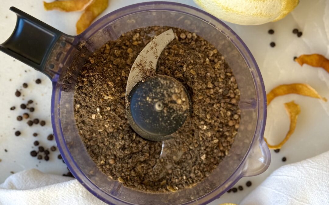 A mini food processor is seen from above, containing Homemade Lemon Pepper Seasoning. The appliance is surrounded by fresh lemons, lemon zest and peppercorns.