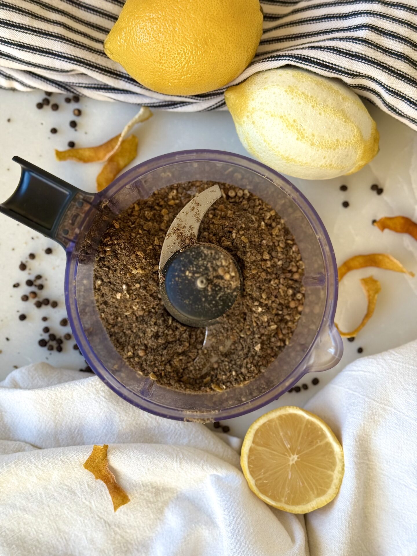 A mini food processor is seen from above, containing Homemade Lemon Pepper Seasoning.  The appliance is surrounded by fresh lemons, lemon zest and peppercorns.