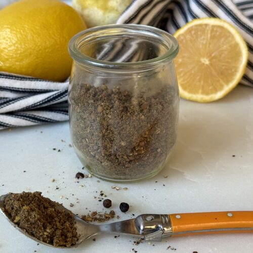 A jar of homemade lemon pepper seasoning sits on a white marble table. A spoonful of the spice blend spills out onto the table in the foreground. Fresh lemons and a black and white striped cloth are seen in the background.