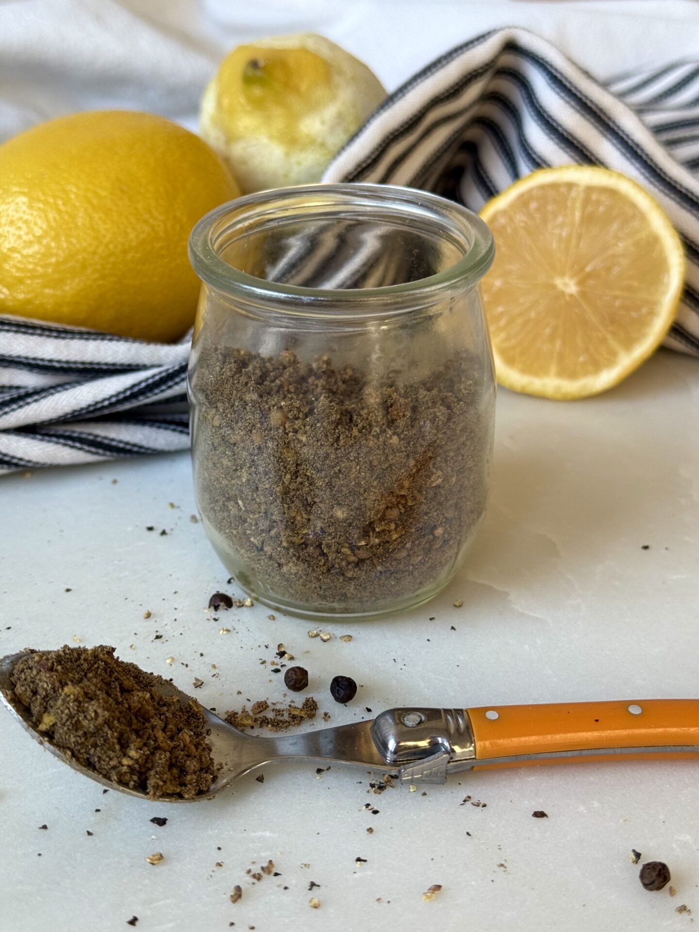 A jar of homemade lemon pepper seasoning sits on a white marble table.  A spoonful of the spice blend spills out onto the table in the foreground.  Fresh lemons and a black and white striped cloth are seen in the background.