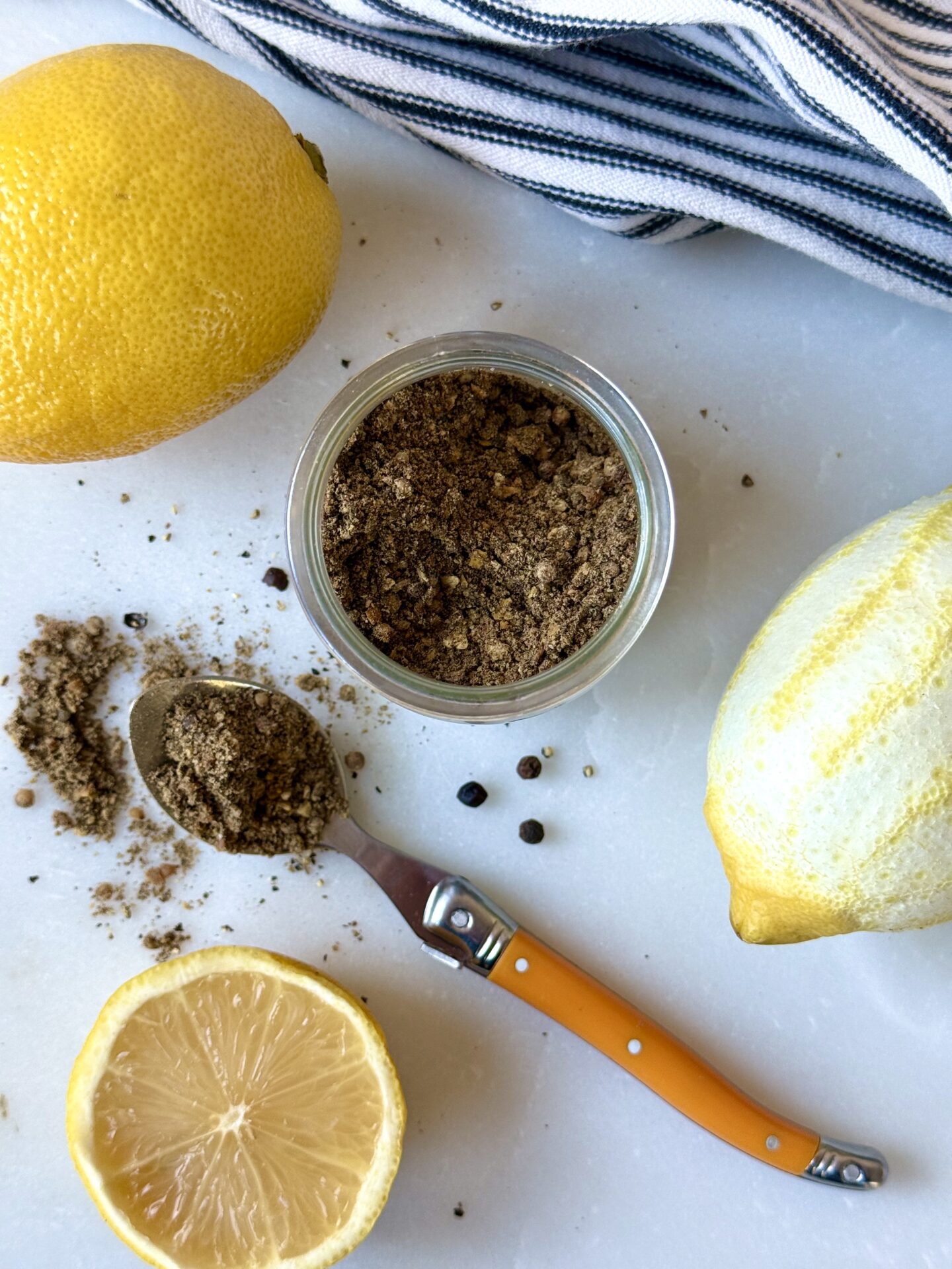 A jar of Homemade Lemon Pepper Seasoning is seen from above, with a spoonfull of the spice mix spilling onto the white marble table.  Fresh lemons and peppercorns surround the image.