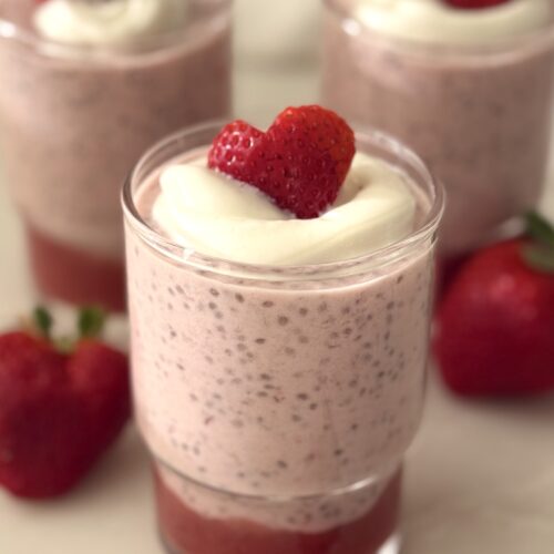 A glass jar holds a Strawberry Chia Seed Pudding with a layer of strawberry puree. The pudding is garnished with a dollop of white yogurt and a heart shaped strawberry slice.