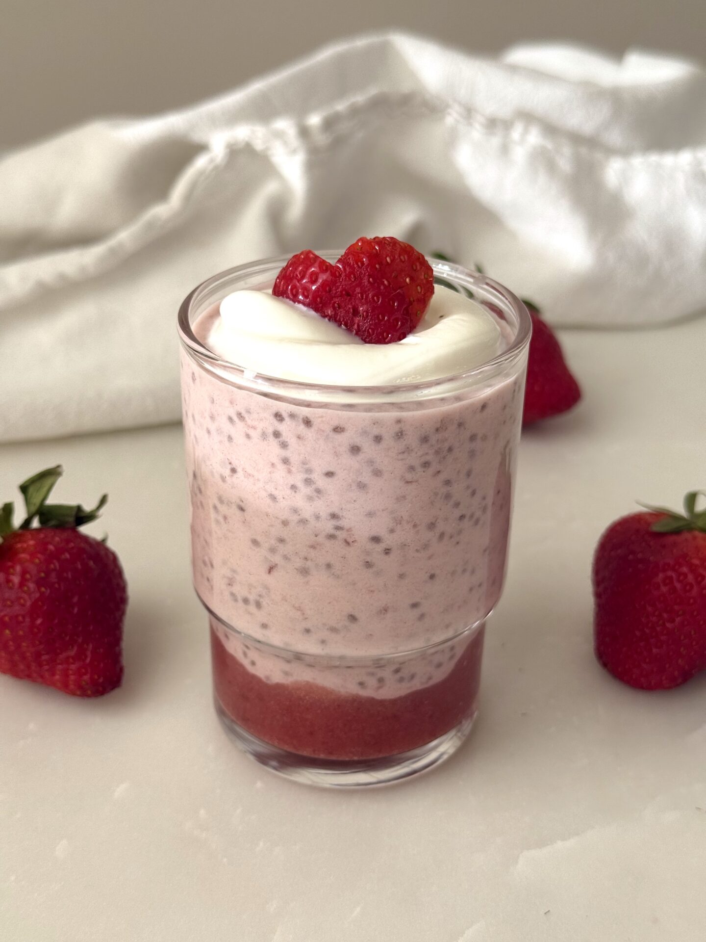 A single serving of strawberry chia seed pudding sits on a white marble table surrounded by fresh strawberries.