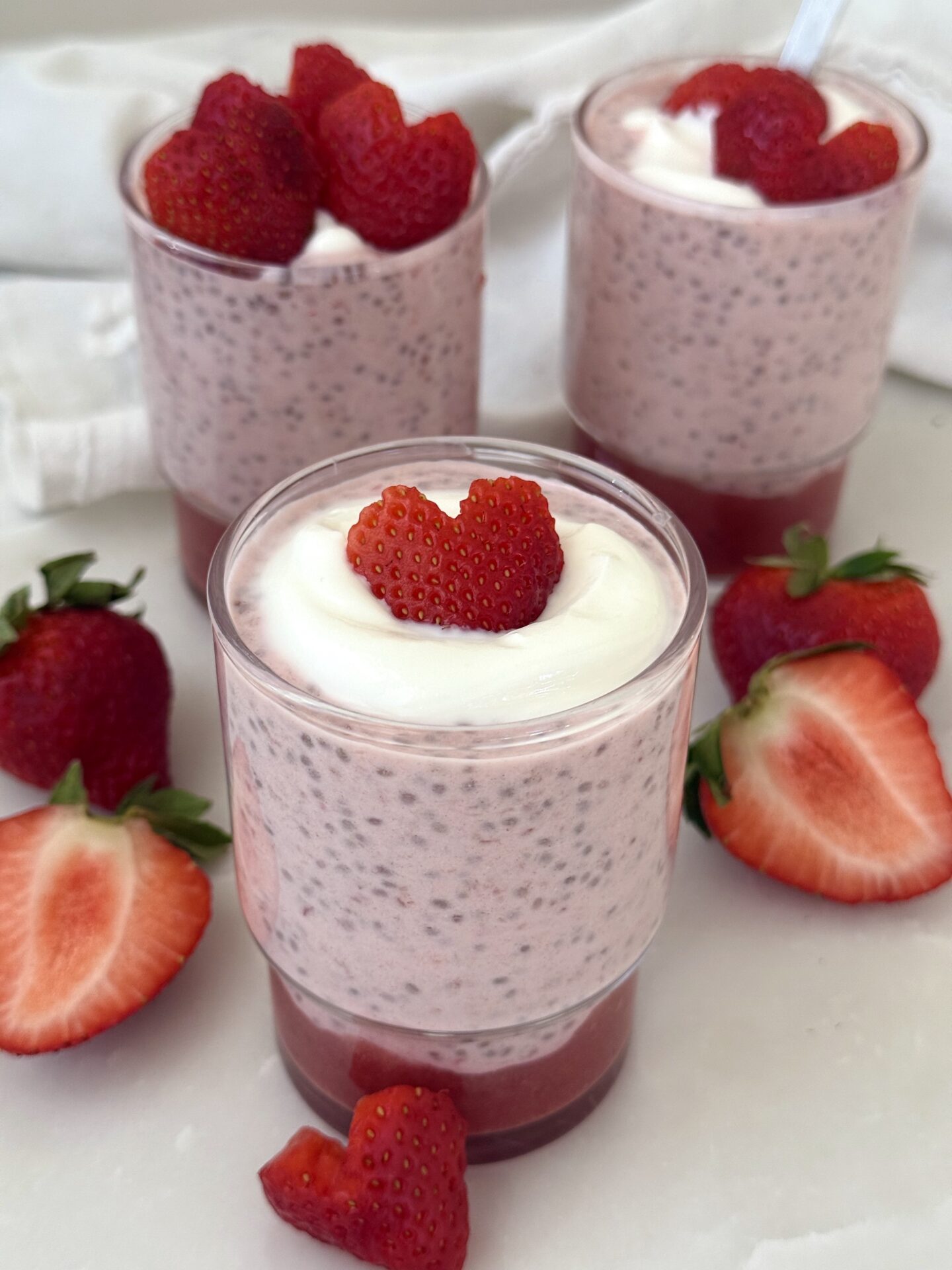 A trio of glass jars hold individual servings of strawberry chia seed pudding, sitting on a white marble table surrounded by fresh strawberries