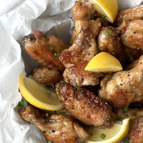 A parchment paper lined bowl of Sweet and Tangy Lemon Pepper Chicken Wings is seen from above, garnished with fresh lemon wedges